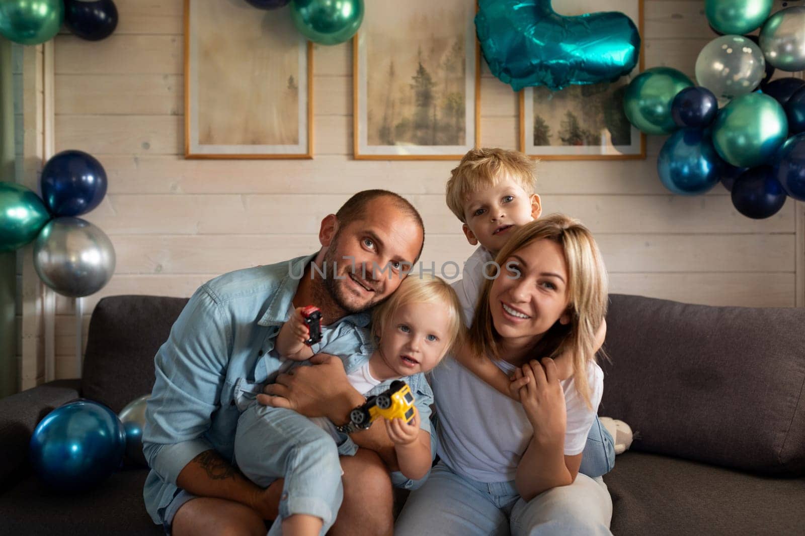 A family of four gathers on a couch, surrounded by colorful balloons. Two children play with toy cars while parents smile, enjoying a joyous moment at home in the afternoon.