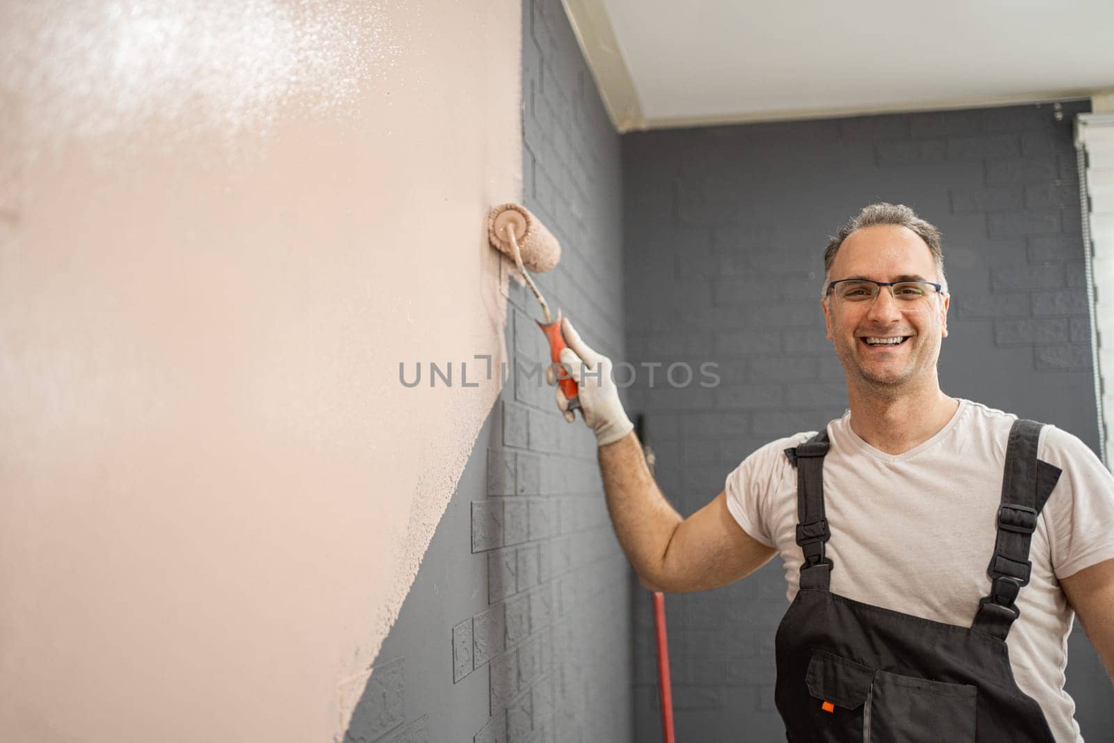 A man is standing in front of a bare wall, actively painting it with a paint roller. He is focused on his task, applying a fresh coat of paint smoothly and evenly.