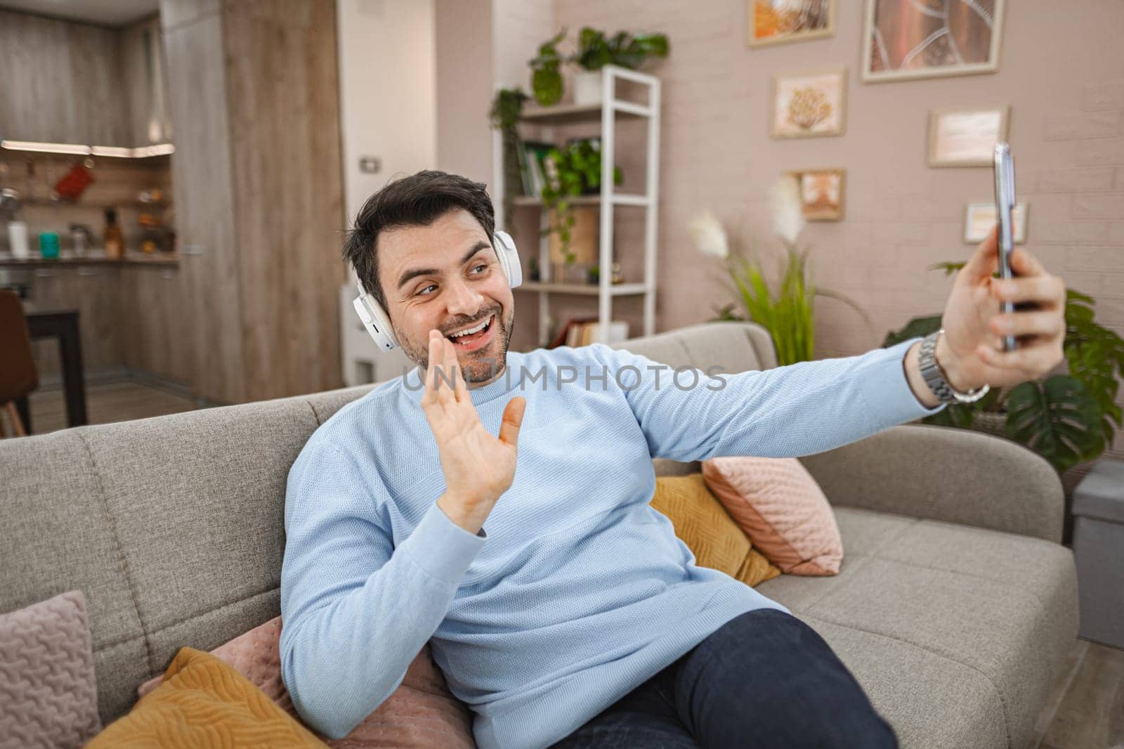 Brown-haired man with white headphones on his head sits on the couch in the living room and makes a video call on a mobile phone.....