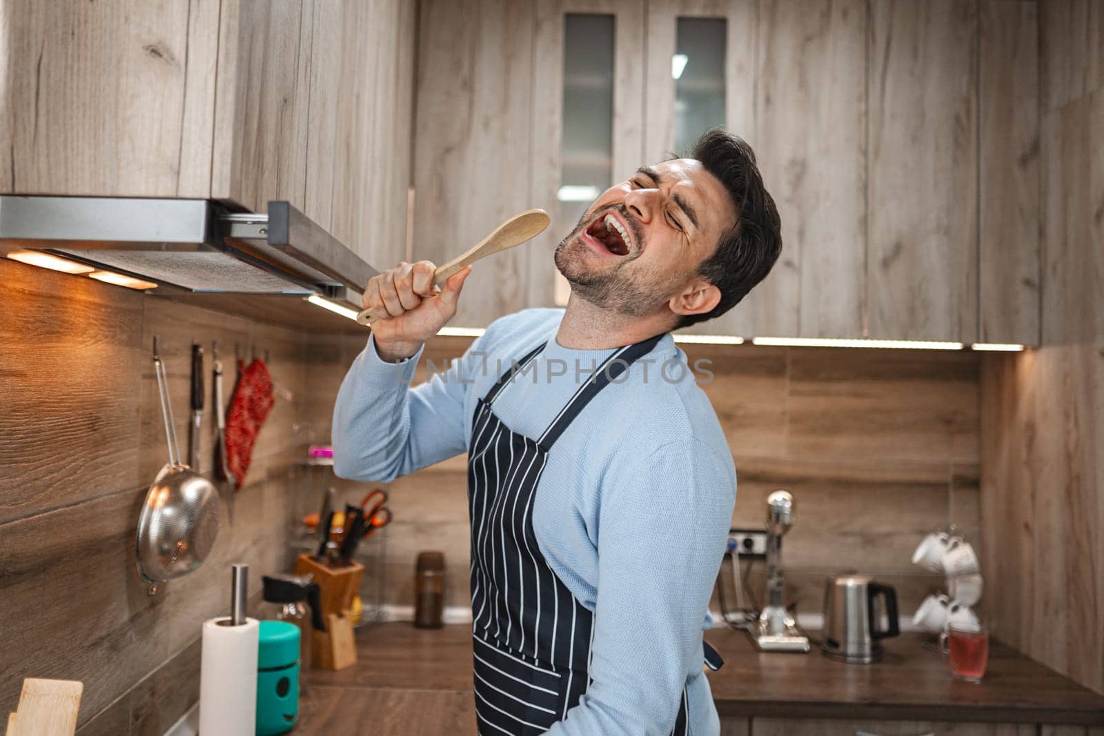 Brown-haired man sings cheerfully into a wooden stove in the kitchen while cooking.