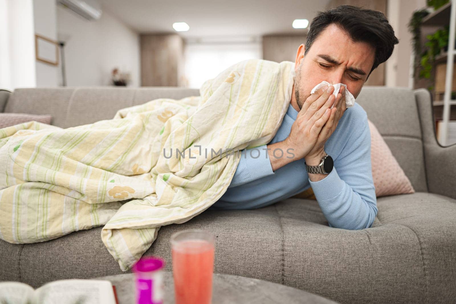 Man is seen laying on a couch, covering his face with a tissue, as he blows his nose due to sickness or allergies. His eyes may appear watery or tired.