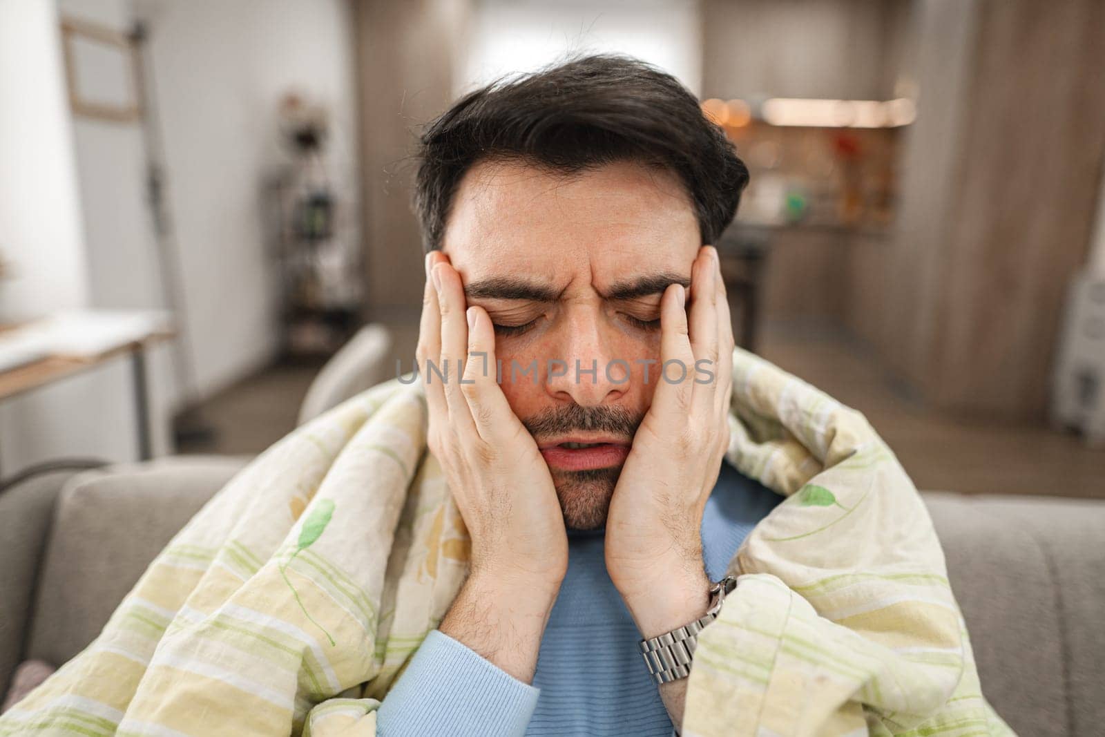 Sick man is sitting covered with a blanket on the sofa in the living room and holding his head with his hands because he has a headache.