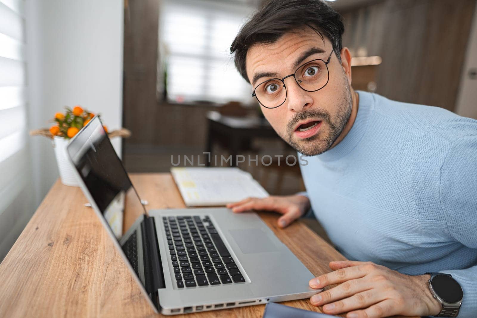 Portrait of a man looking shocked at the camera, sitting at a desk in the living room and working on a laptop