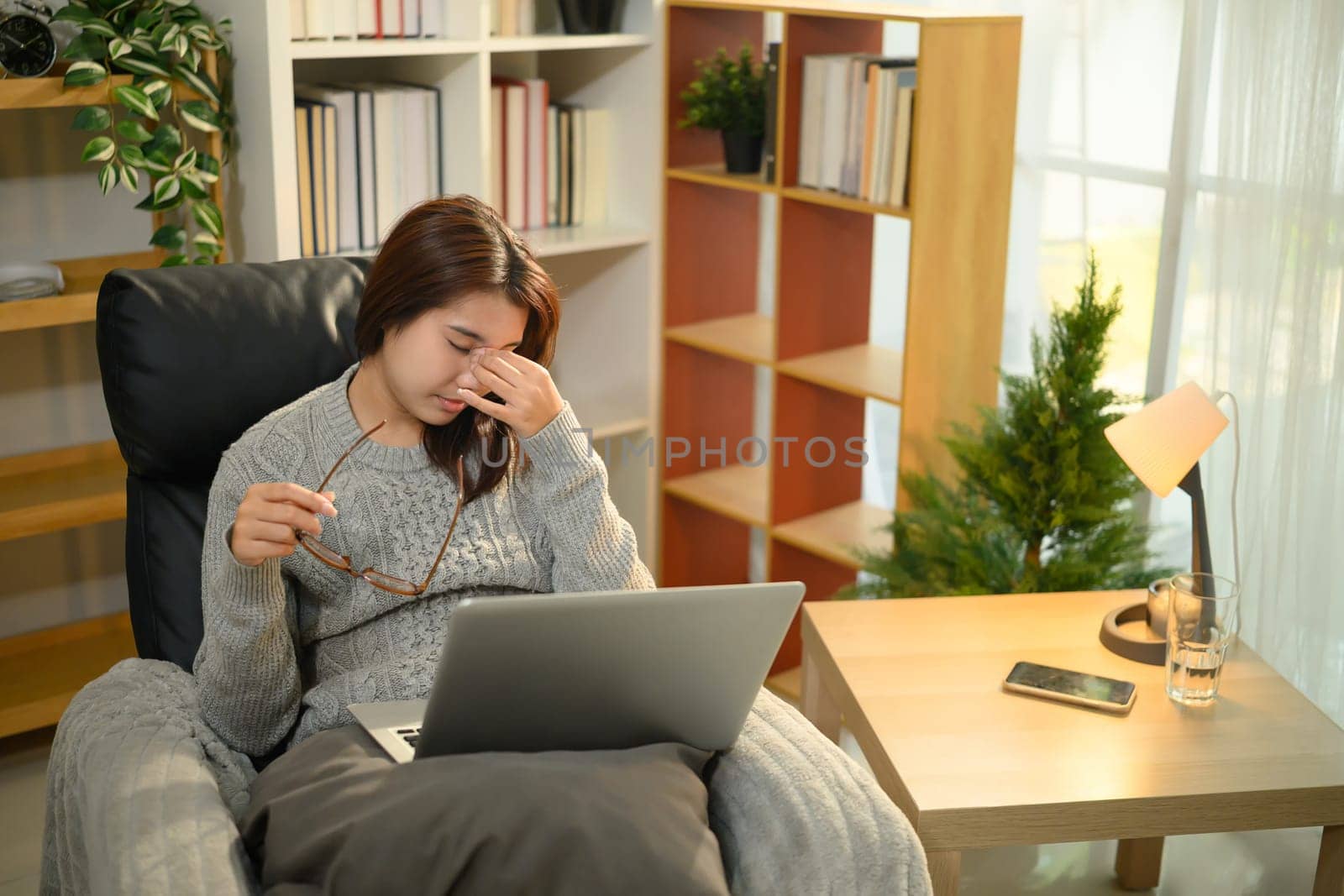 Tired woman removes glasses and rubs her eyes from fatigue while working on laptop at home.