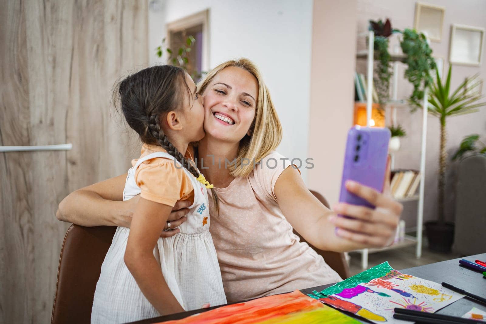 Daughter kisses her mother as they happily capture a selfie together in their kitchen.