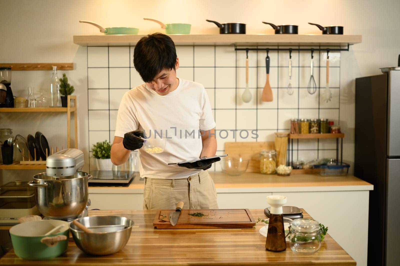 Smiling asian man follows an online recipe on digital tablet while preparing whipped cream.