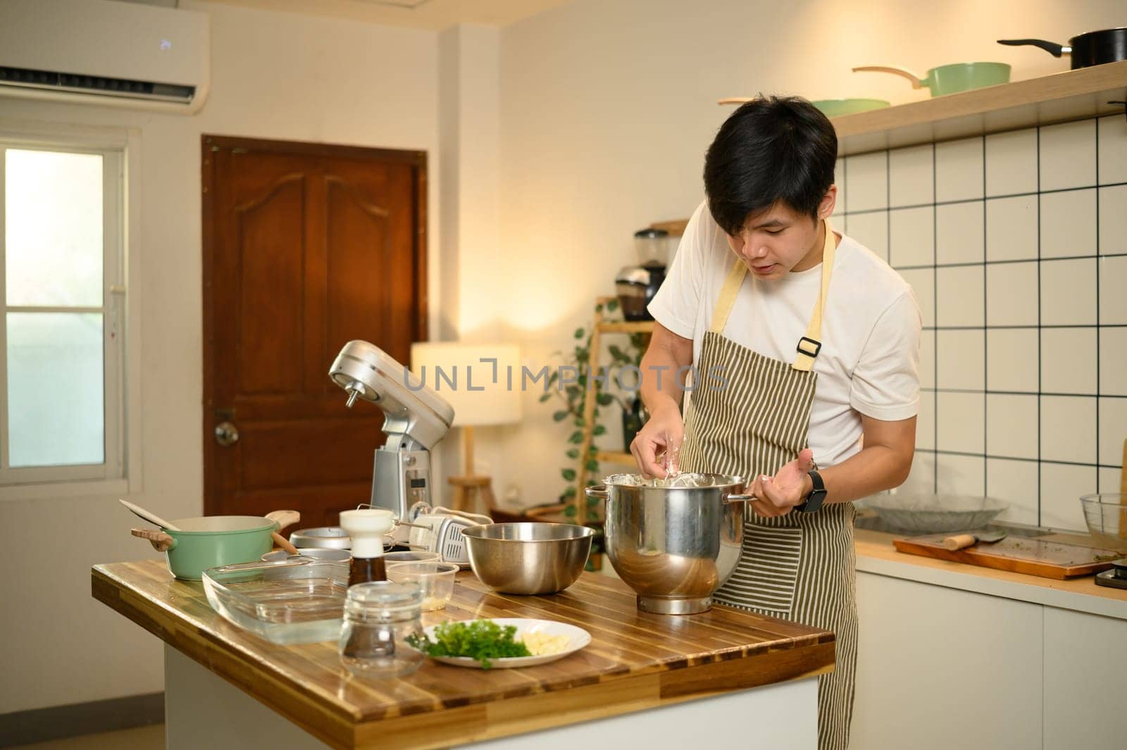 Young asian man preparing whipped cream in stainless steel bowl, using whisk in a bright kitchen.