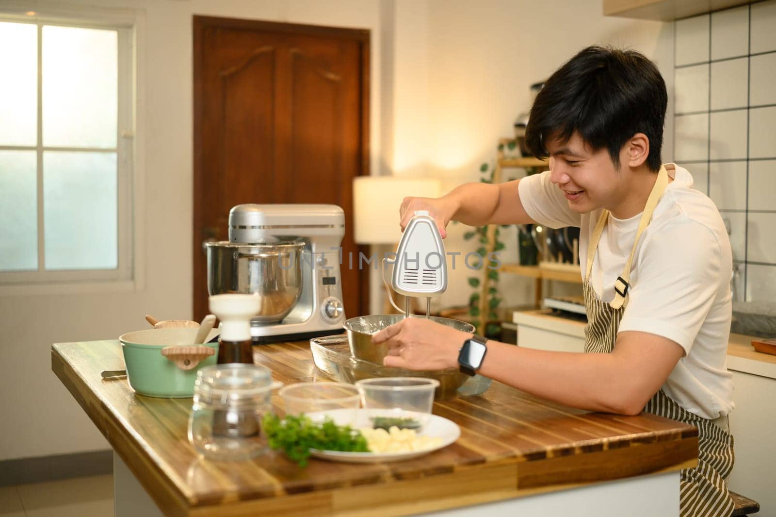 Young man in apron, blending cream with hand mixer in a stainless steel bowl.