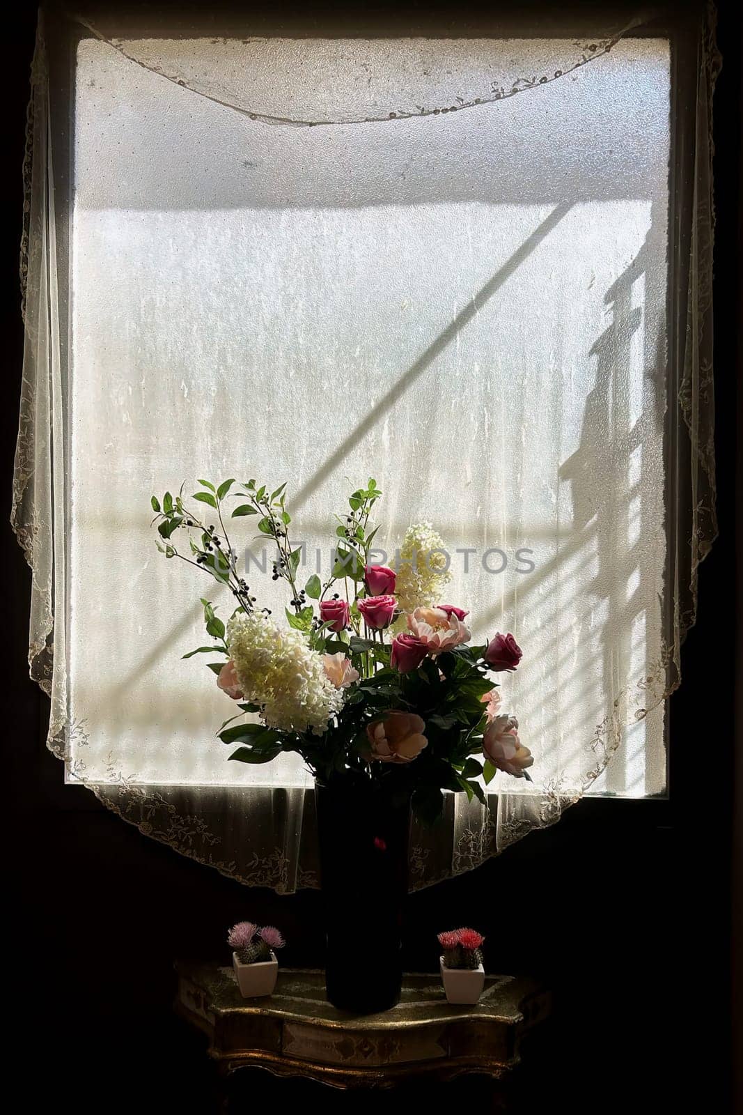 A captivating still life photograph featuring a lush bouquet of flowers in a dark vase, positioned against a window draped with sheer, lace-trimmed curtains. The soft, diffused light streaming through the window illuminates the flowers, creating a serene and elegant composition. This image evokes a sense of tranquility and delicate beauty, perfect for projects requiring a touch of sophistication and natural charm.