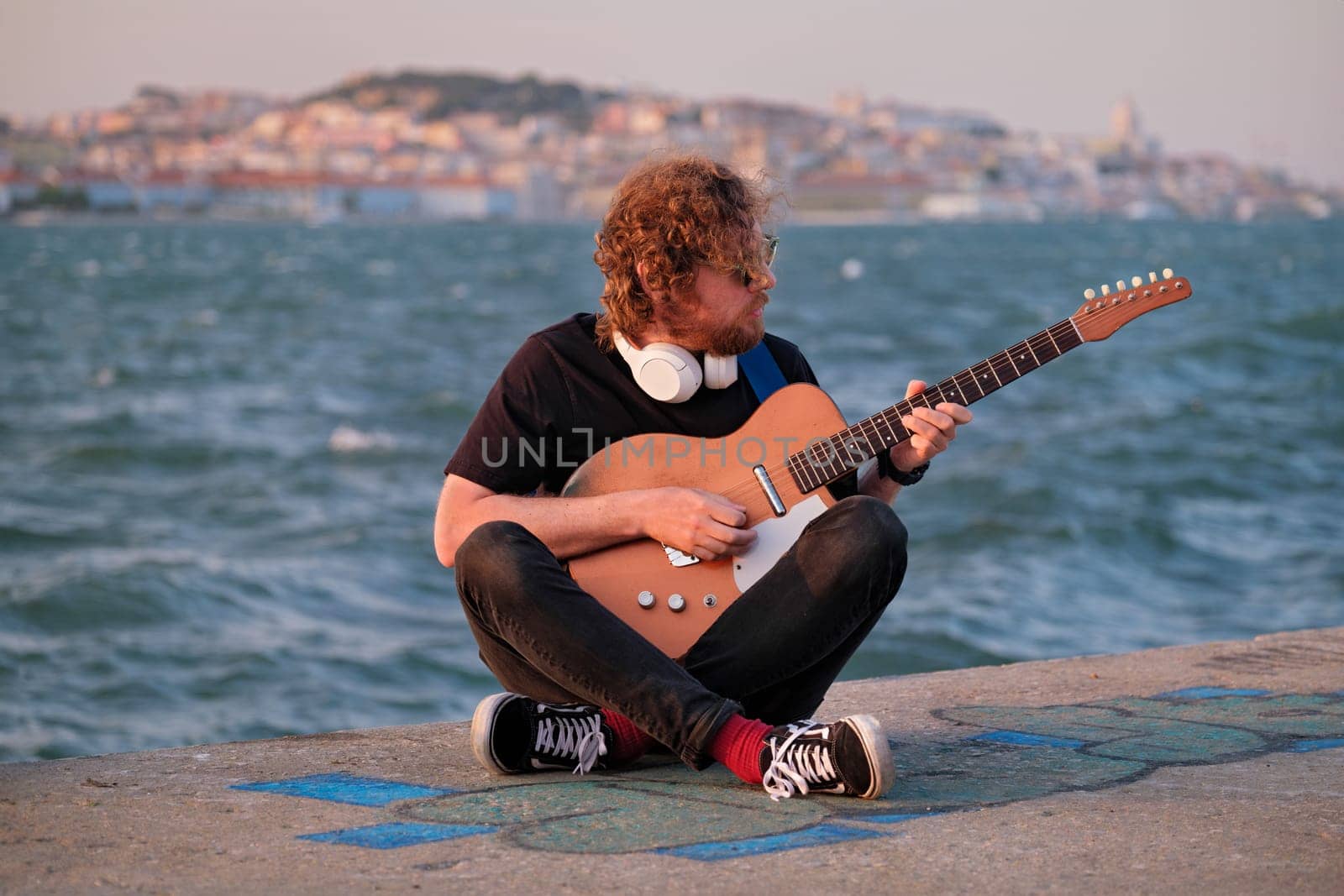Hipster street musician in black playing electric guitar in the street on sunset on embankment with Lisbon in background. Portugal