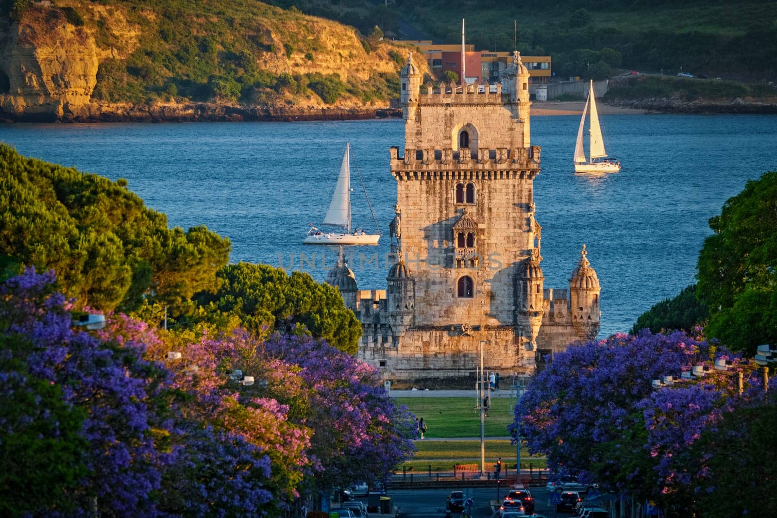 Scenic view of Belem Tower in Lisbon, Portugal, seen over a street with blooming purple jacaranda flower trees street with tourist sailboats on the Tagus River on sunset. Portugal