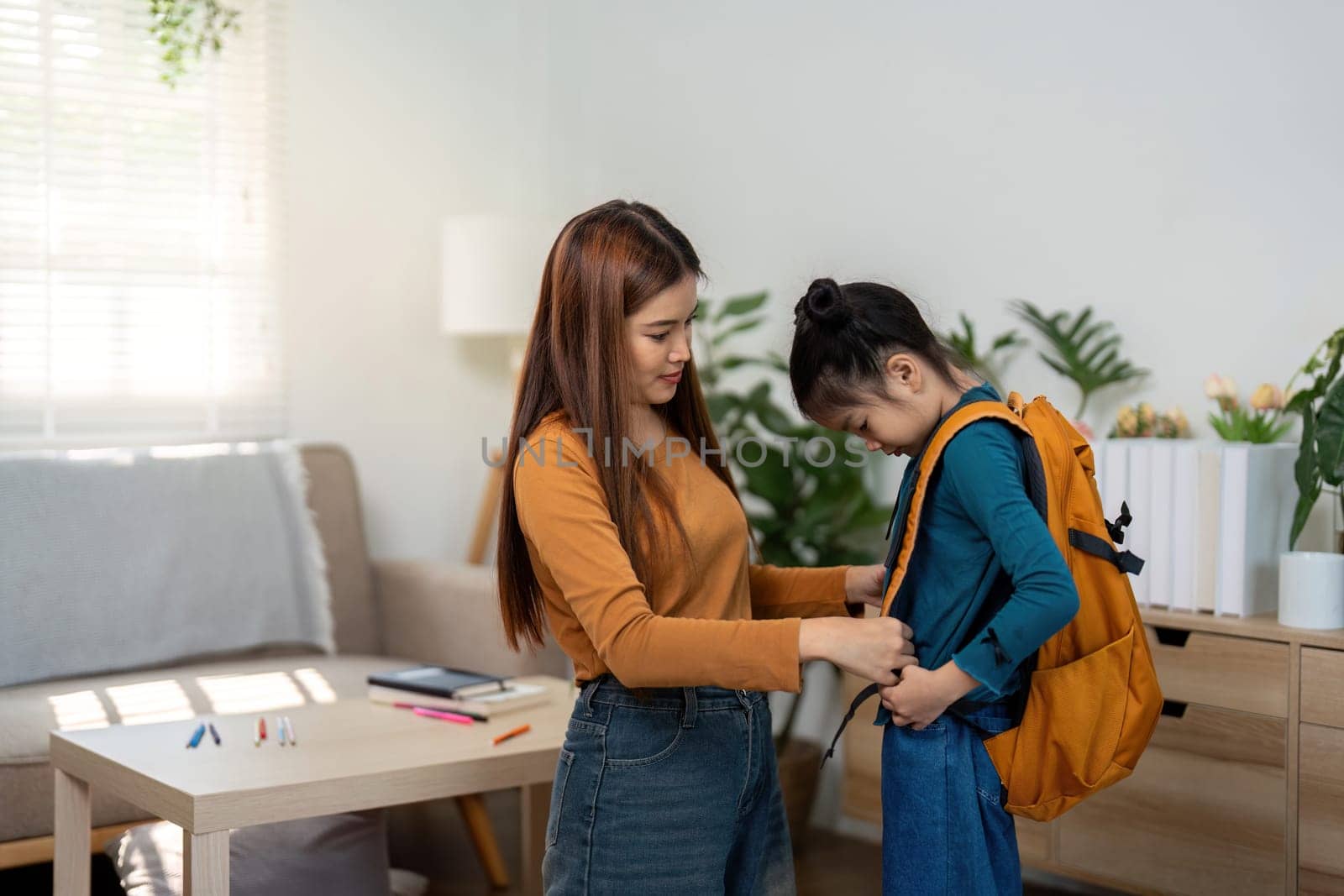 The mother assists her daughter in putting on her backpack, preparing her for a day at school, showcasing nurturing parenting.