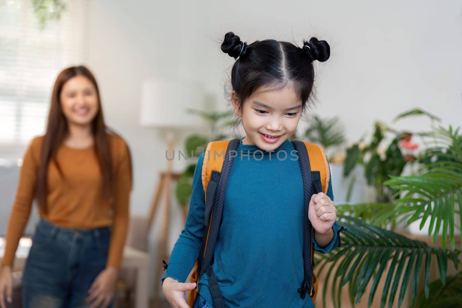A cheerful young girl wears a bright backpack as she walks towards the door, ready for school. Her smiling mother observes, highlighting the warmth of family support.