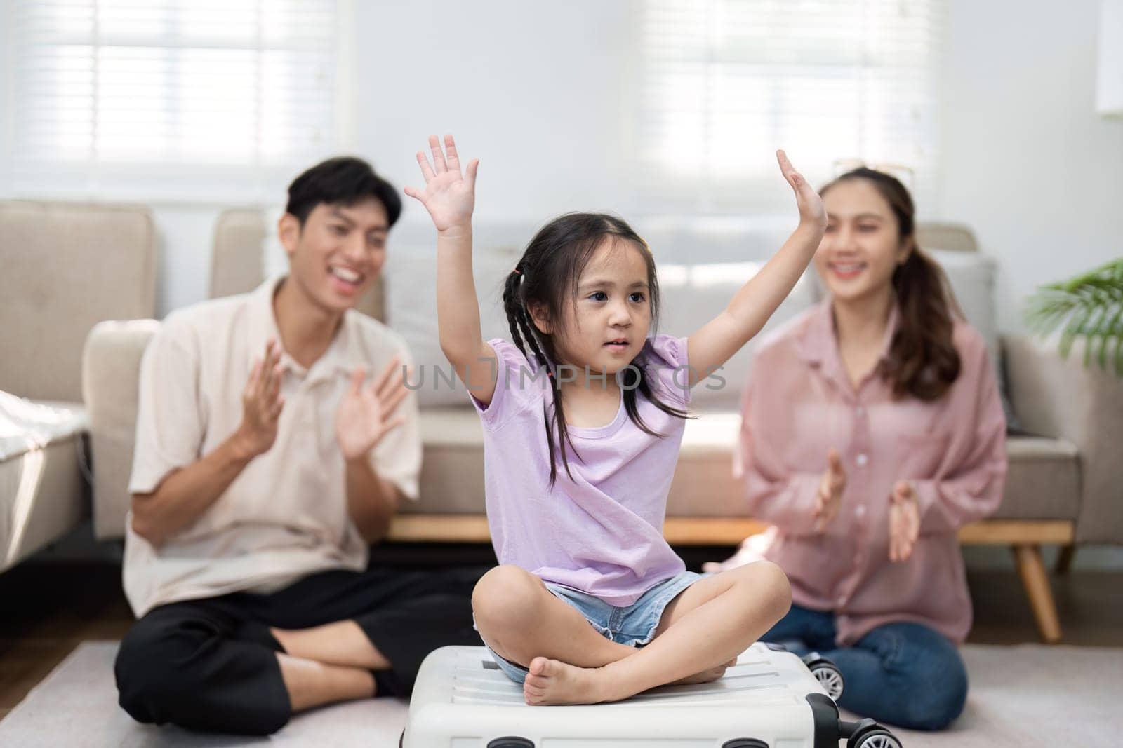 In a lighthearted moment, a girl raises her hands in excitement while sitting on a suitcase, as her joyful parents celebrate the fun of preparing for their family journey.
