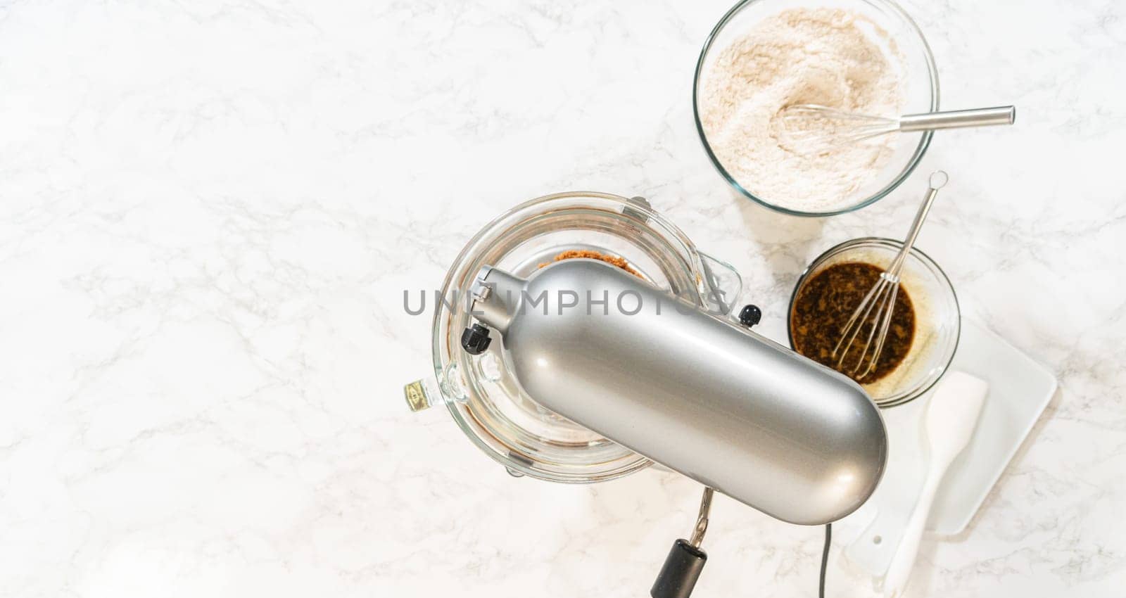 Overhead view of a baking setup for Classic Gingerbread Cookie Dough, with a stand mixer surrounded by essential ingredients. Ingredients include brown sugar, butter, flour mixture, and dark molasses in individual glass bowls, showcasing the classic components needed for this festive dough recipe.