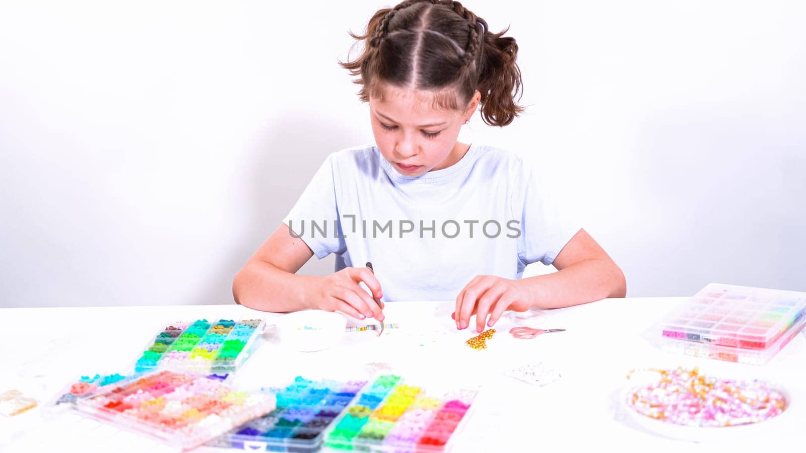 Delicate fingers of a young girl navigate through a treasure trove of bright, multicolored beads, each compartment revealing a new hue to choose from. She's immersed in the joyful task of stringing together a handmade bracelet, with golden beads and pearls lying nearby to add a touch of sparkle to her creation.