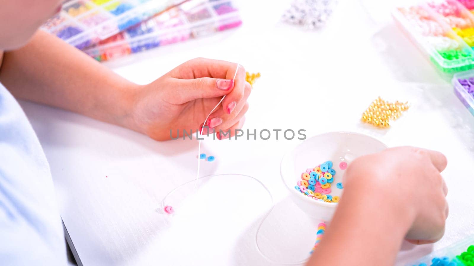 Delicate fingers of a young girl navigate through a treasure trove of bright, multicolored beads, each compartment revealing a new hue to choose from. She's immersed in the joyful task of stringing together a handmade bracelet, with golden beads and pearls lying nearby to add a touch of sparkle to her creation.