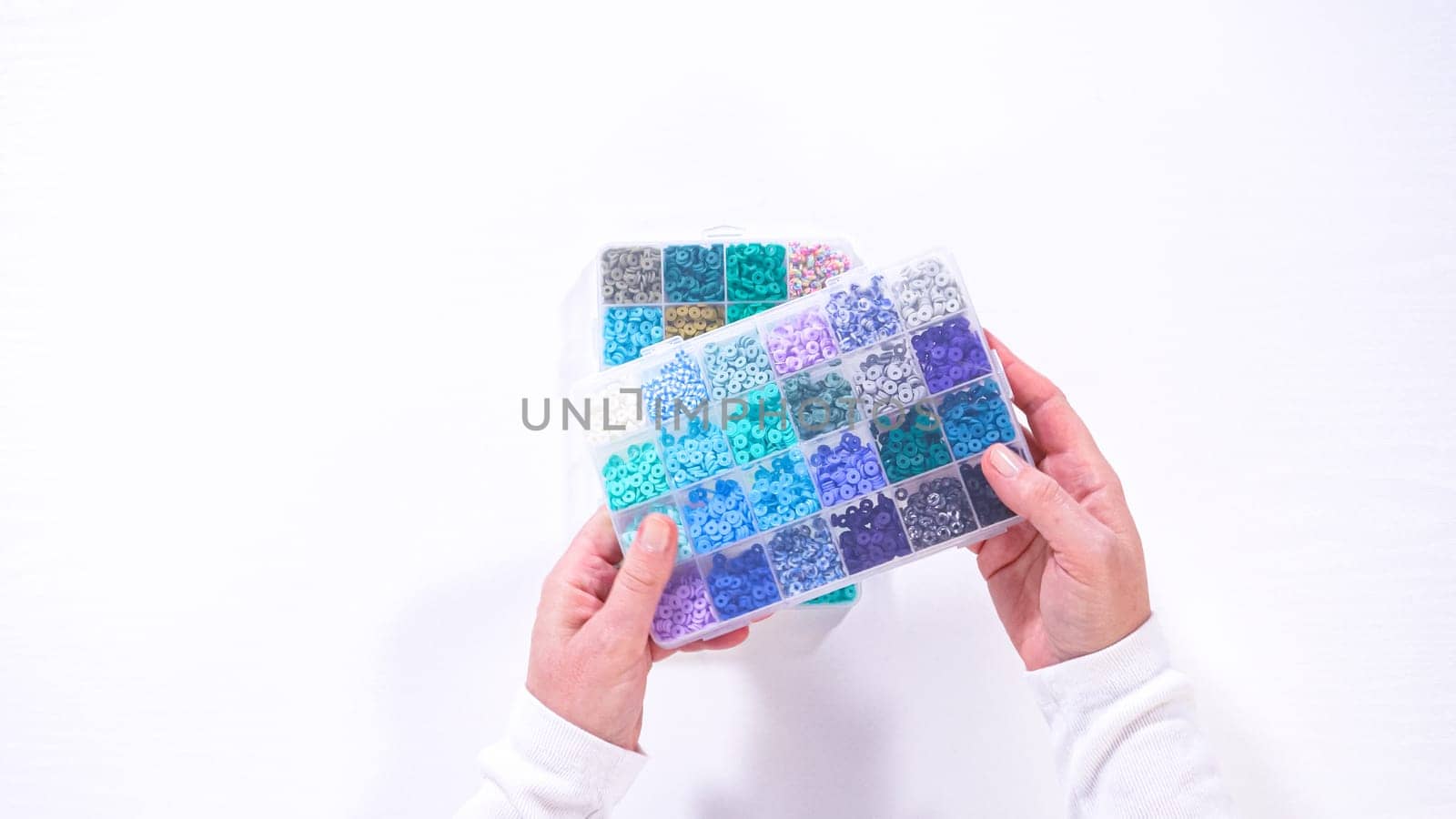 Flat lay. Woman hands gracefully poised over a collection of beads, sorted by color in transparent organizers. The array of beads spans a vibrant spectrum, from deep purples to bright oranges, meticulously arranged for easy selection as she embarks on creating a custom piece of jewelry.