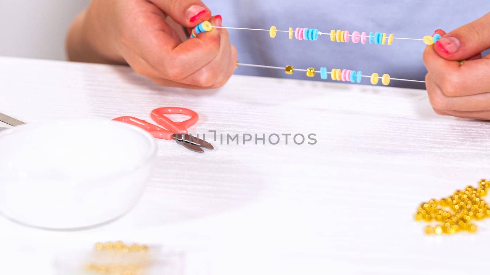 Delicate fingers of a young girl navigate through a treasure trove of bright, multicolored beads, each compartment revealing a new hue to choose from. She's immersed in the joyful task of stringing together a handmade bracelet, with golden beads and pearls lying nearby to add a touch of sparkle to her creation.