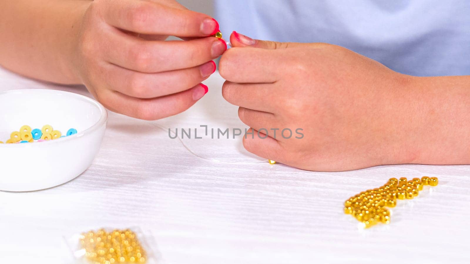 Delicate fingers of a young girl navigate through a treasure trove of bright, multicolored beads, each compartment revealing a new hue to choose from. She immersed in the joyful task of stringing together a handmade bracelet, with golden beads and pearls lying nearby to add a touch of sparkle to her creation.