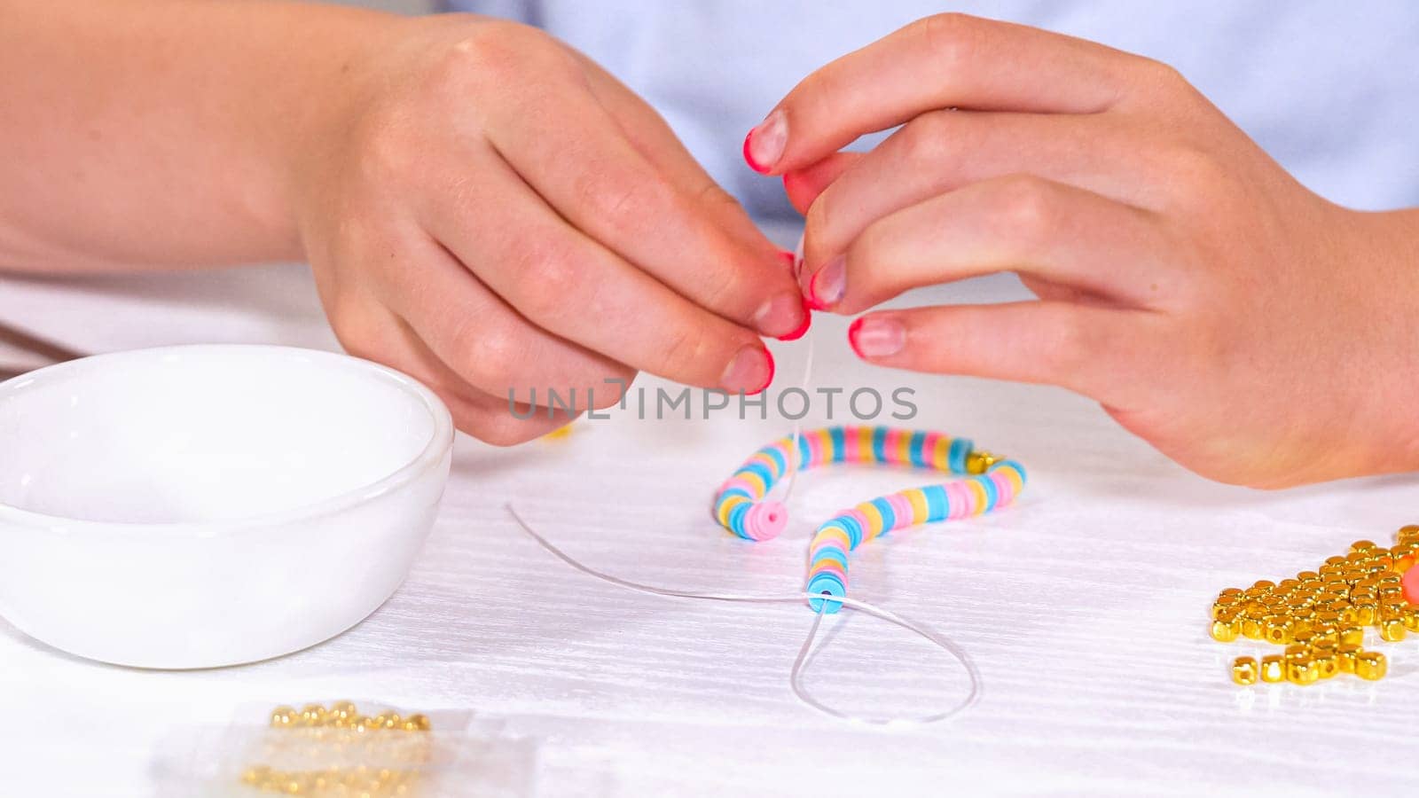 Delicate fingers of a young girl navigate through a treasure trove of bright, multicolored beads, each compartment revealing a new hue to choose from. She's immersed in the joyful task of stringing together a handmade bracelet, with golden beads and pearls lying nearby to add a touch of sparkle to her creation.