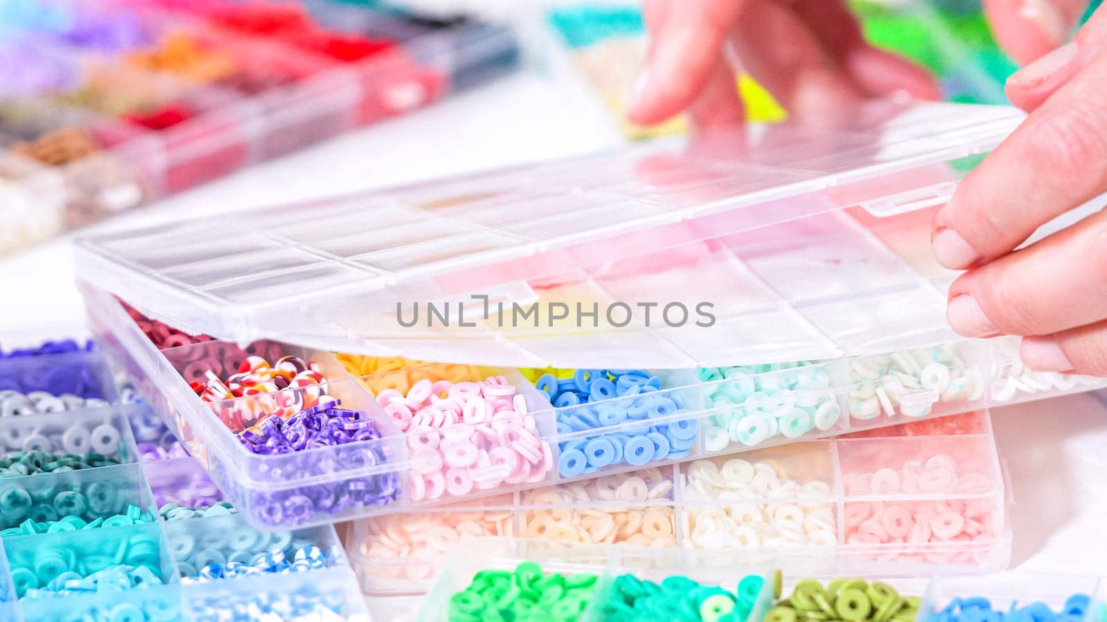 Woman hands gracefully poised over a collection of beads, sorted by color in transparent organizers. The array of beads spans a vibrant spectrum, from deep purples to bright oranges, meticulously arranged for easy selection as she embarks on creating a custom piece of jewelry.