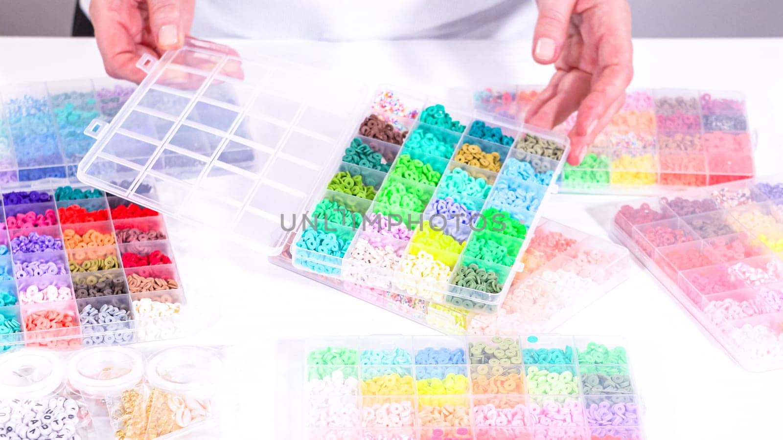 Woman hands gracefully poised over a collection of beads, sorted by color in transparent organizers. The array of beads spans a vibrant spectrum, from deep purples to bright oranges, meticulously arranged for easy selection as she embarks on creating a custom piece of jewelry.