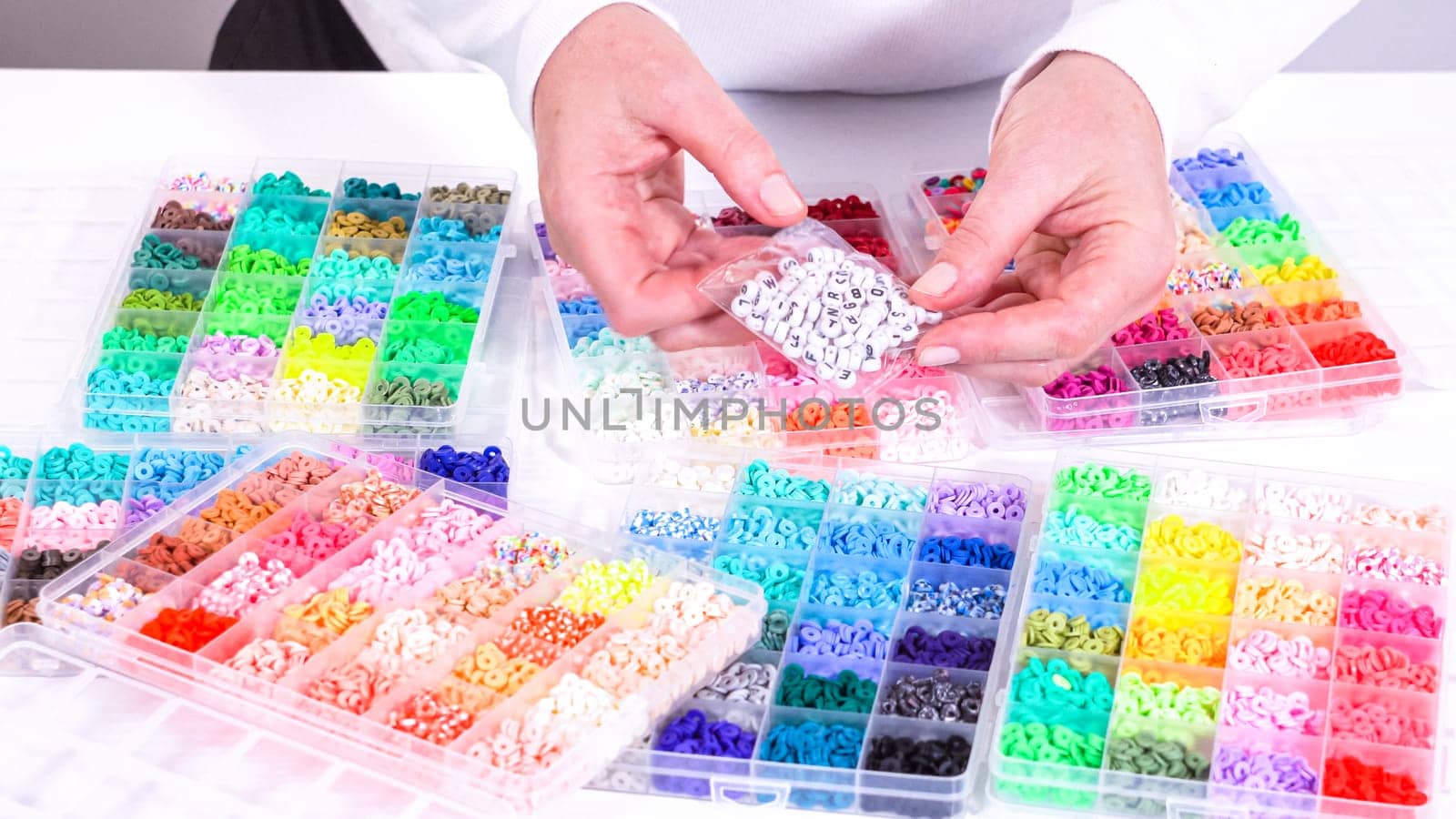 Woman hands gracefully poised over a collection of beads, sorted by color in transparent organizers. The array of beads spans a vibrant spectrum, from deep purples to bright oranges, meticulously arranged for easy selection as she embarks on creating a custom piece of jewelry.