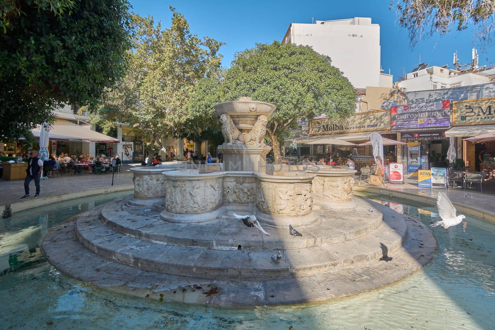 Crete.Greece - april 07, 2025: Magnificent Morosini fountain featuring four intricately carved lions spouting water, surrounded by vibrant greenery and bustling activity in Heraklion's historic plaza.
