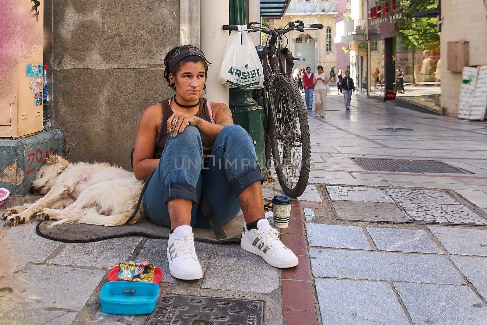 Crete.Greece - april 07, 2025: A homeless individual seated on the street with a loyal white dog resting nearby. Surrounding objects include a bicycle, small container, and coffee cup, highlighting urban survival.