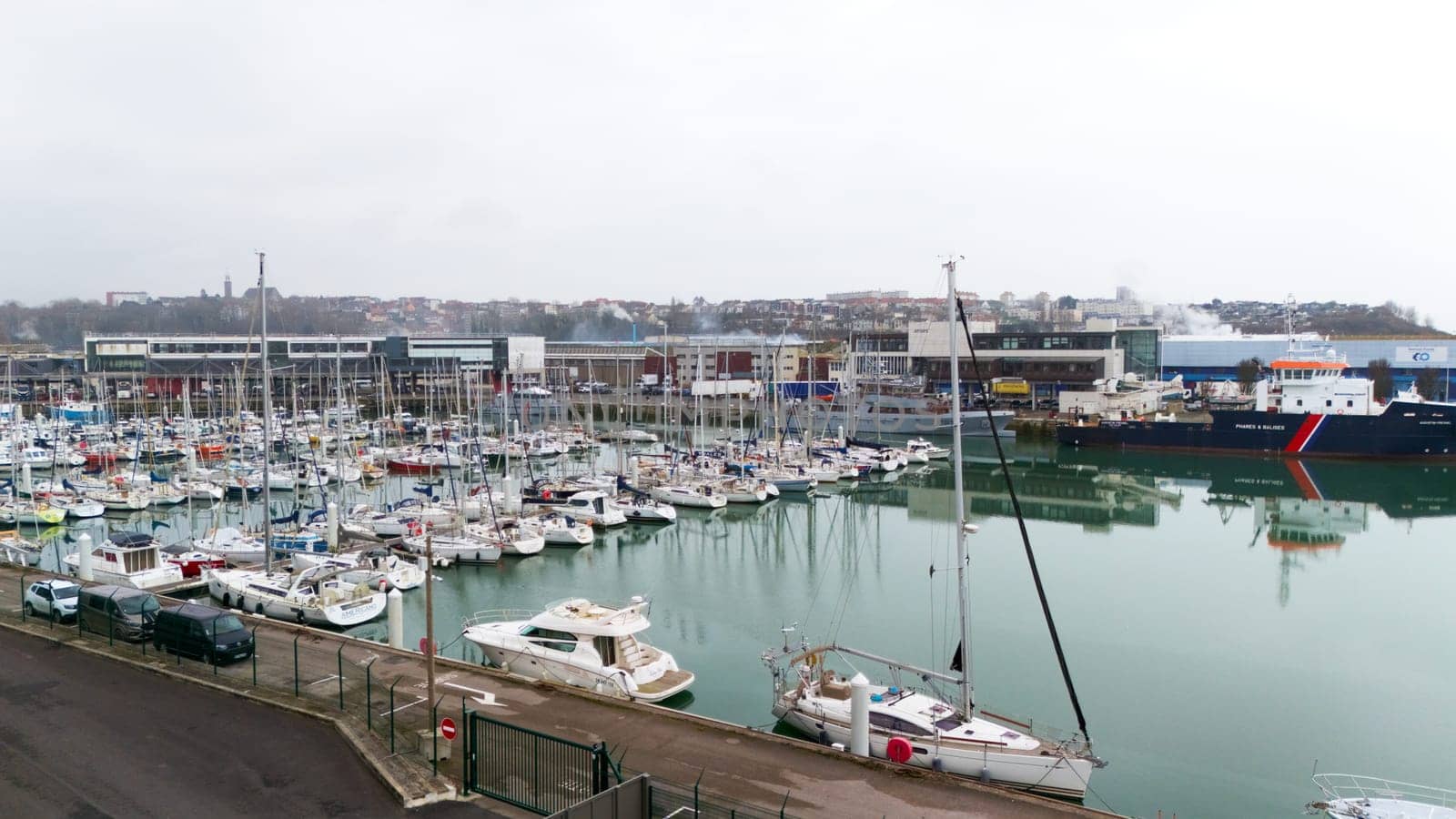 Boulogne. France 03.28.2025 Overcast view of a marina filled with sailboats and yachts, bordered by fencing and industrial buildings in the background.