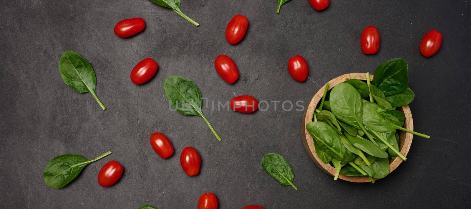 A wooden bowl filled with fresh spinach leaves surrounded by scattered cherry tomatoes and spinach on a dark background, top view