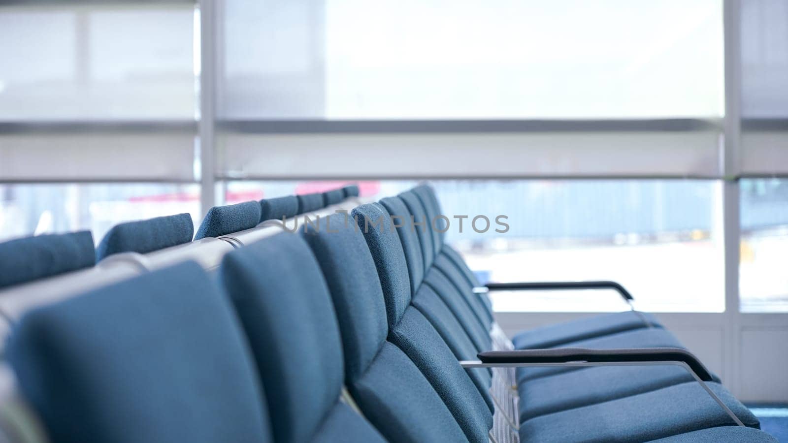 This is a photo of a row of empty blue airport seats with a large glass window in the background. The photo is taken from the perspective of a passenger sitting in one of the seats.