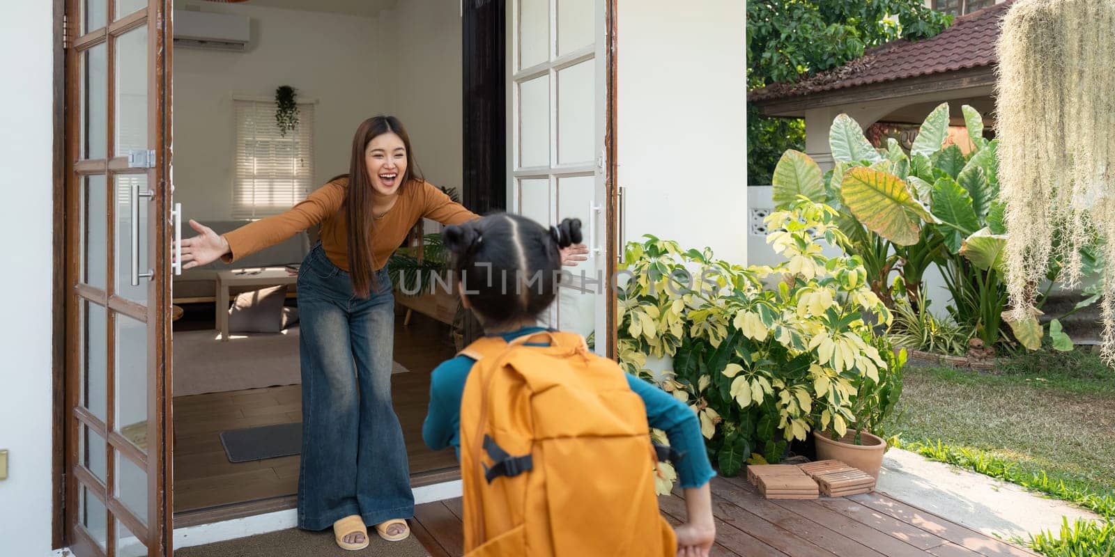 A cheerful mother opens her arms wide to greet her daughter, who is returning home from school, highlighting the joy of family togetherness.