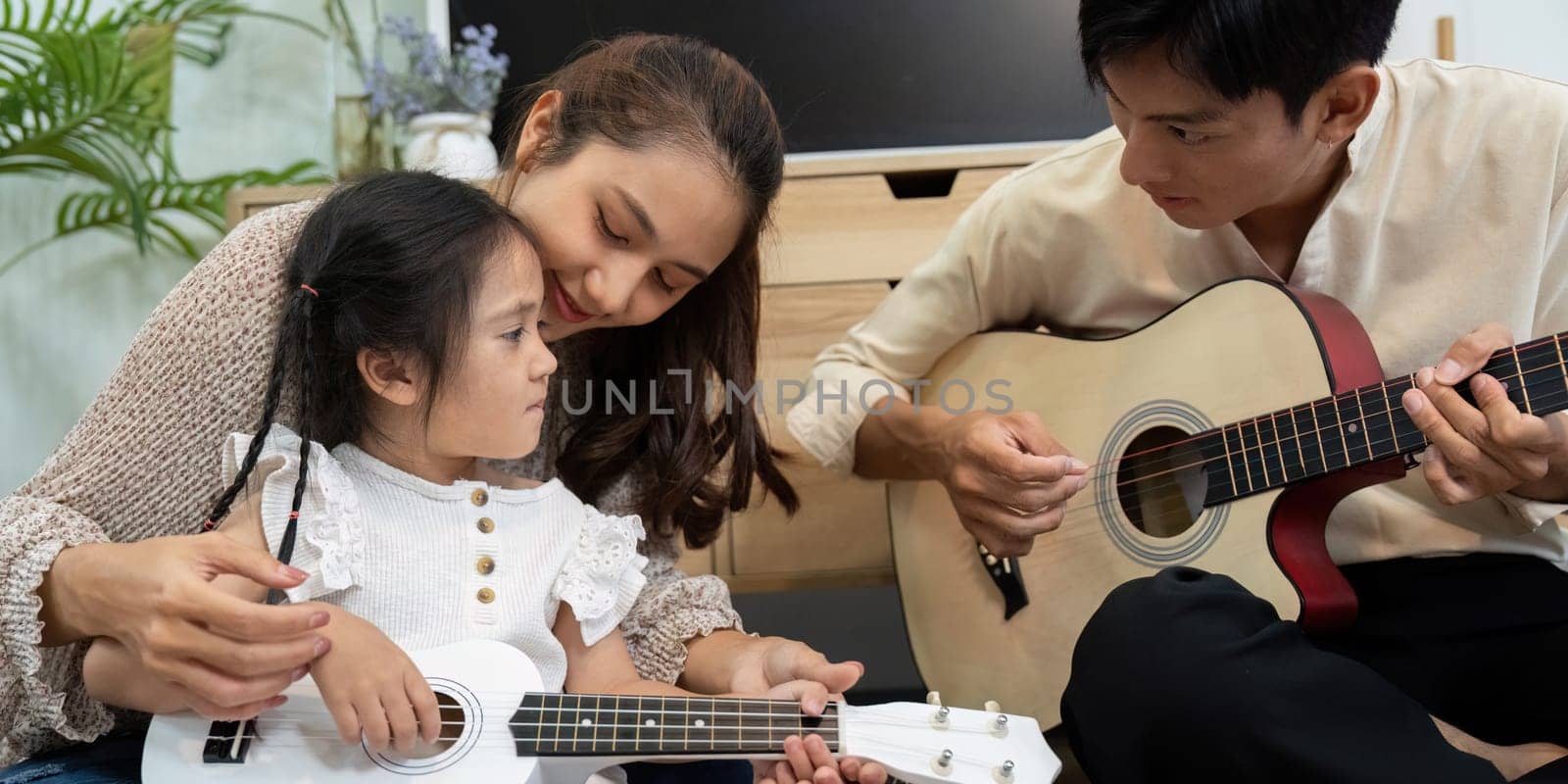 In a cozy living room, a mother lovingly teaches her daughter to play the ukulele. Their interaction emphasizes the joy of family bonding and musical learning.