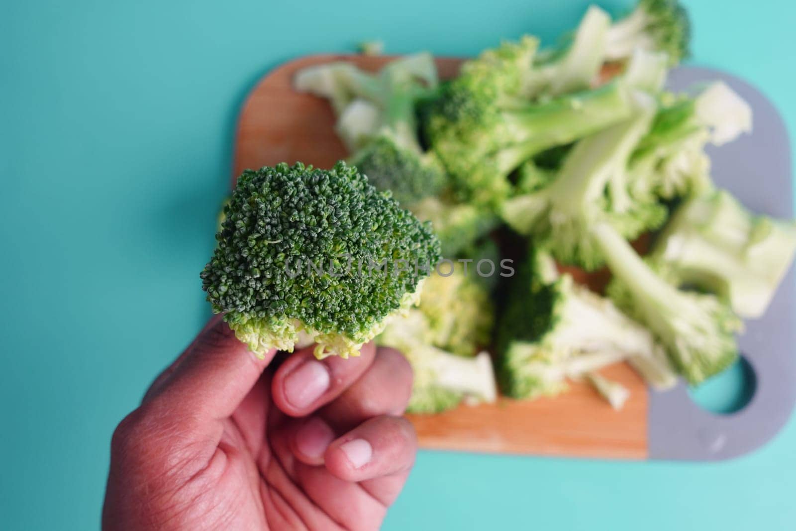 Broccoli floret is held above a wooden cutting board with more broccoli.