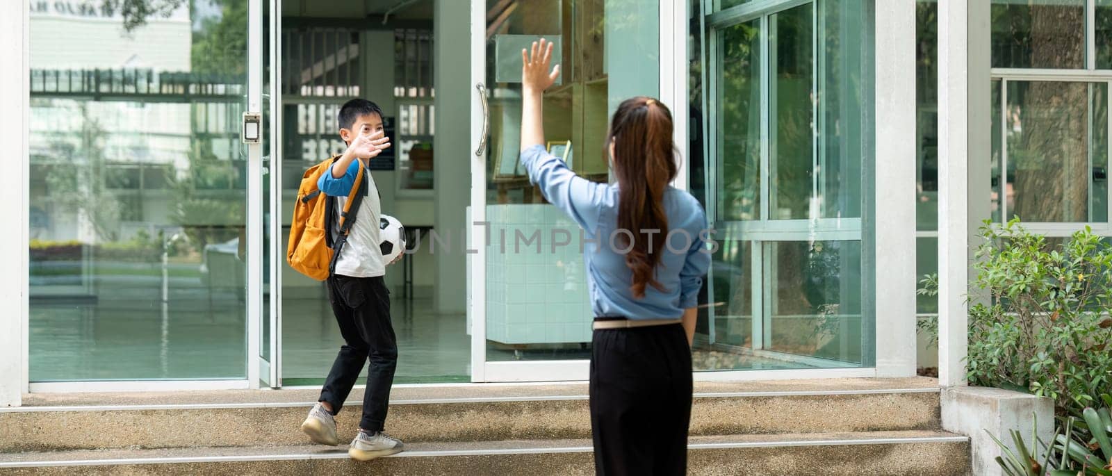 The young boy waves back to his mother while entering school, showcasing the bond and support between them, and the excitement of school life.