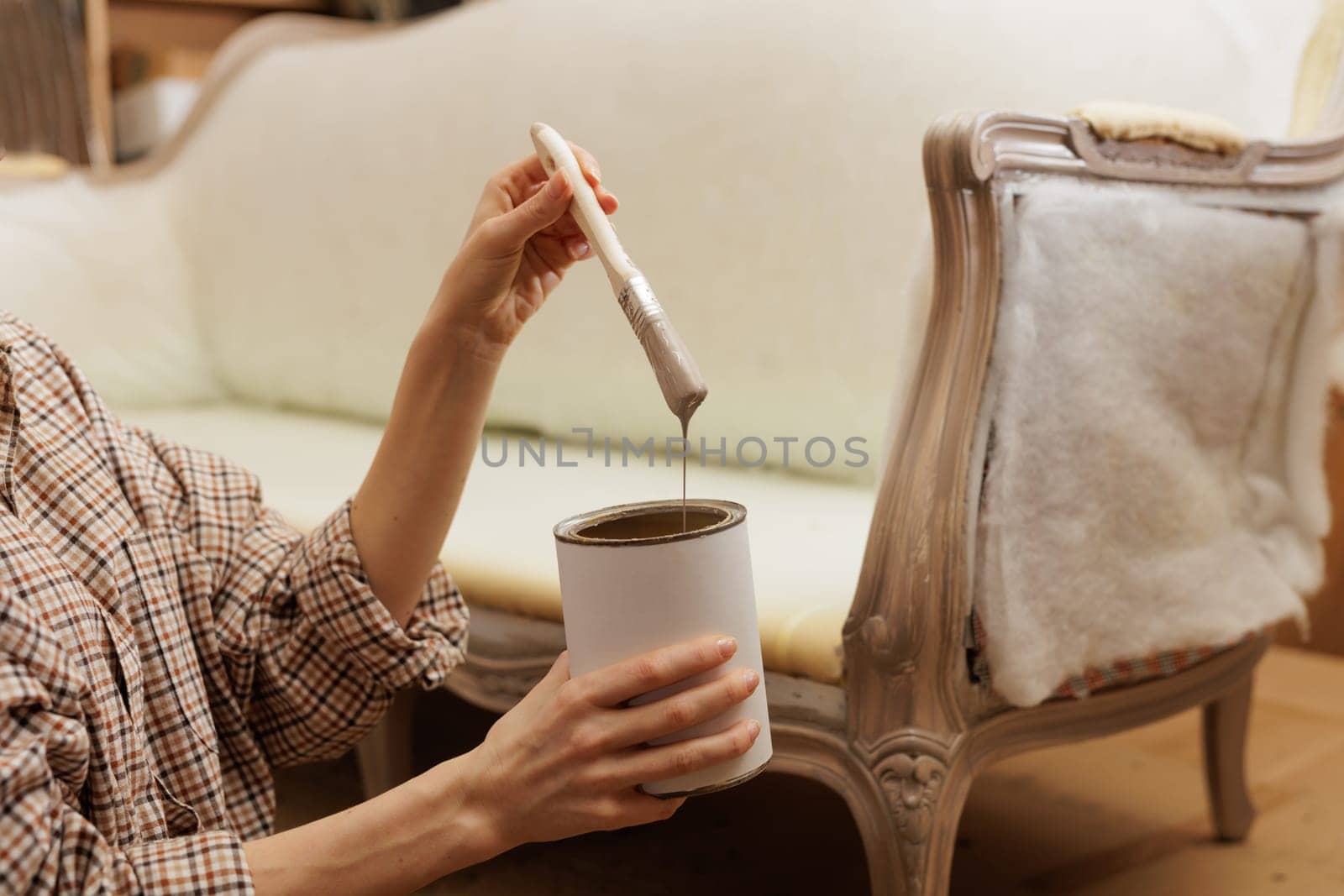 Close-up of woman's hands holding a paintbrush dripping with beige paint, restoring vintage furniture at home.