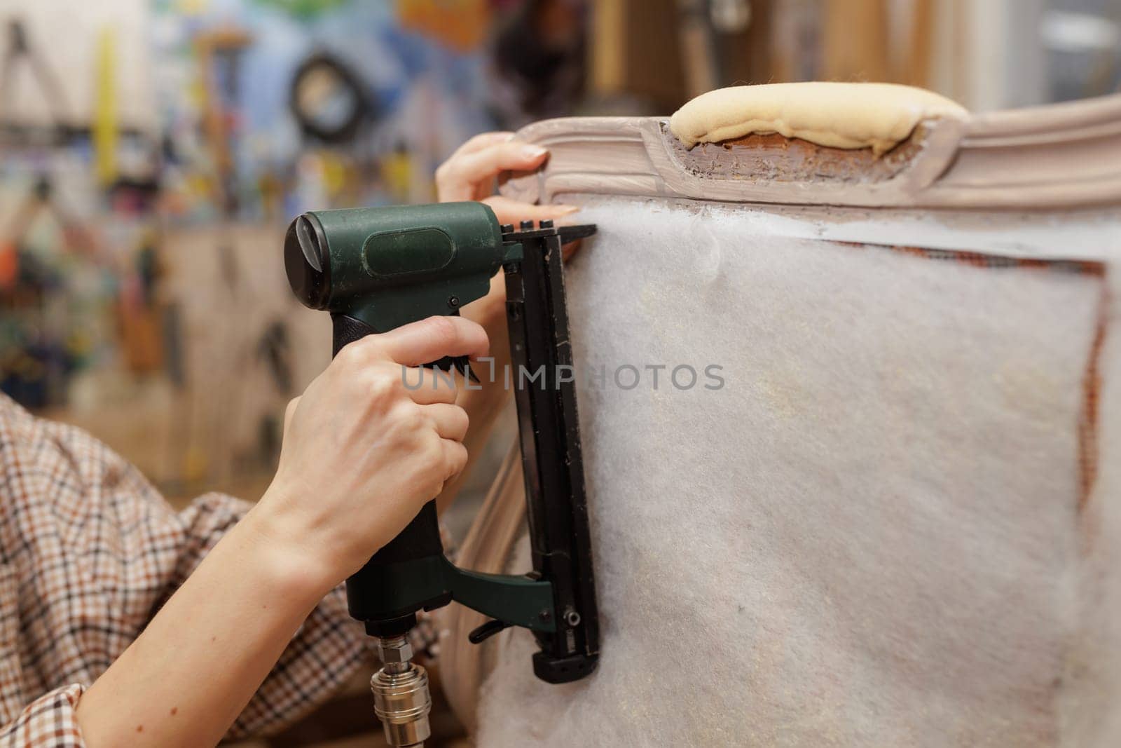 Close-up of hands using a pneumatic stapler to attach padding on a vintage armchair during upholstery work.