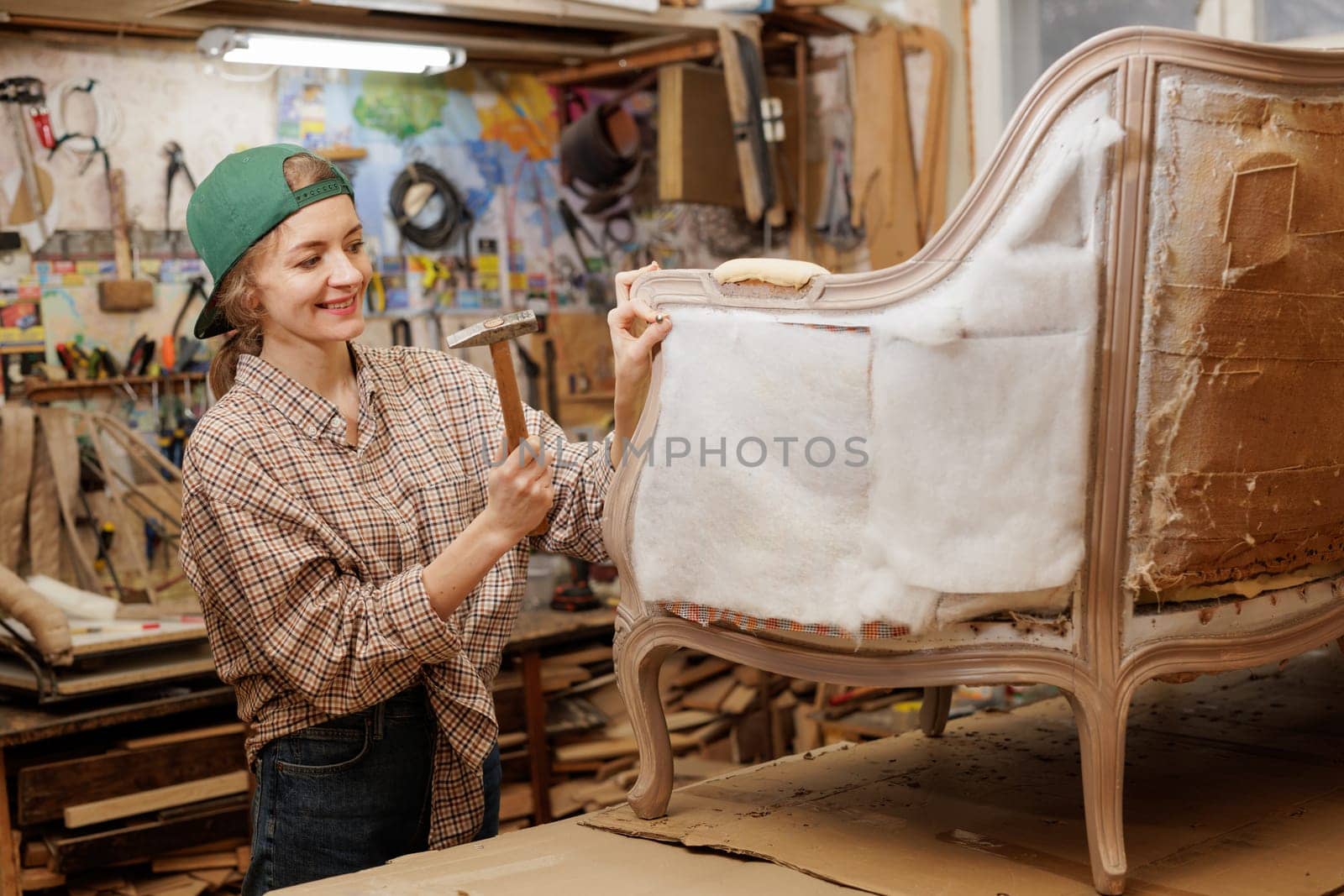 Happy female artisan upholstering furniture in workshop, repairing vintage armchair, using hammer and smiling, creative upholstery work concept.