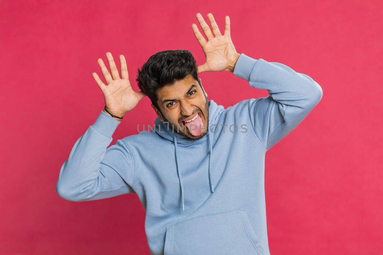 Funny comical playful Indian man making silly facial expressions, grimacing, fooling around, showing tongue, idiotic expression. Comedian Arabian guy isolated on red background. People lifestyles