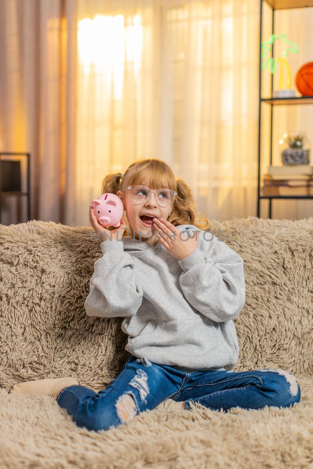 Young teenager girl holds a toy piggy bank at home, rattling it gently to hear the metallic coins clink. Child kid on sofa smiles broadly, thrilled excited about setting save money for a dream item.