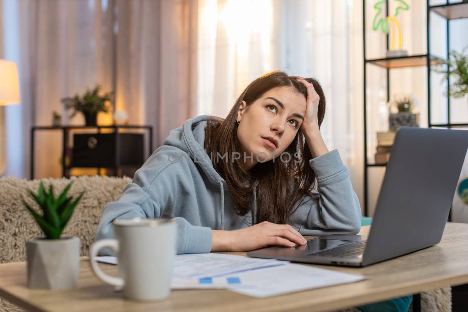 Exhausted lazy young girl freelancer working on laptop at table on sofa looking tired at the end of workday holding head with one hand. Documents, cup of coffee are on table. Woman working from home