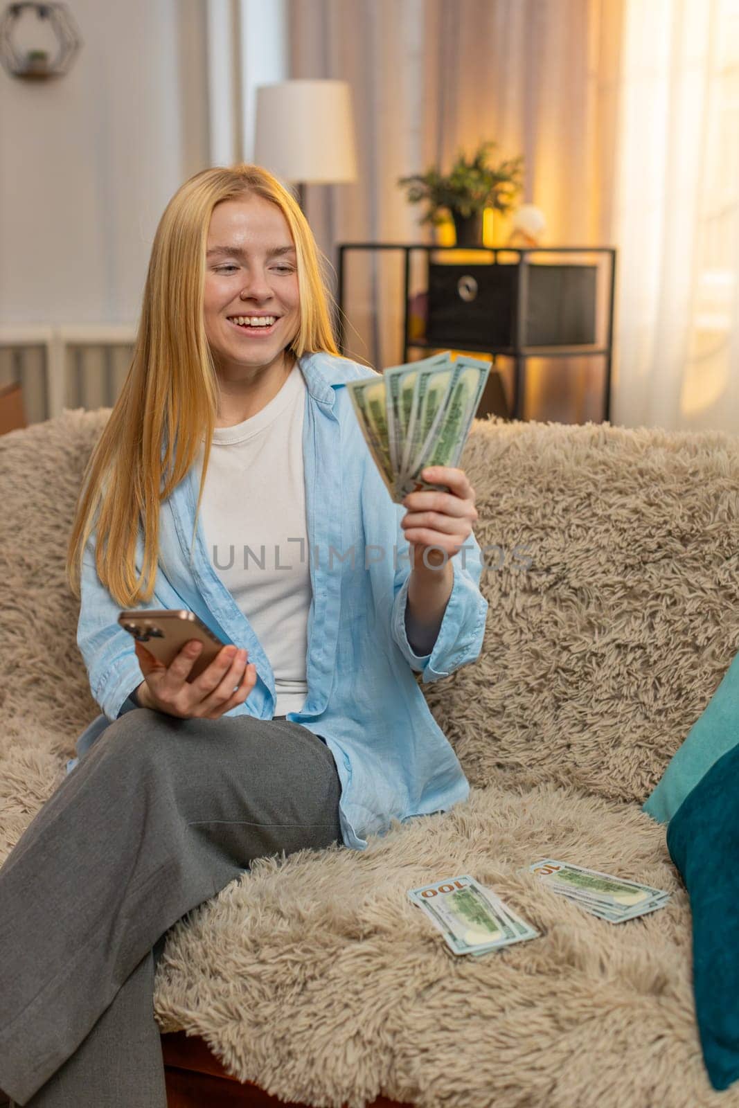 Young woman relaxes at home, counting banknotes while entering sums on smartphone to find extra savings. Caucasian girl on sofa celebrates with triumph pose, feeling proud of unexpected financial gain