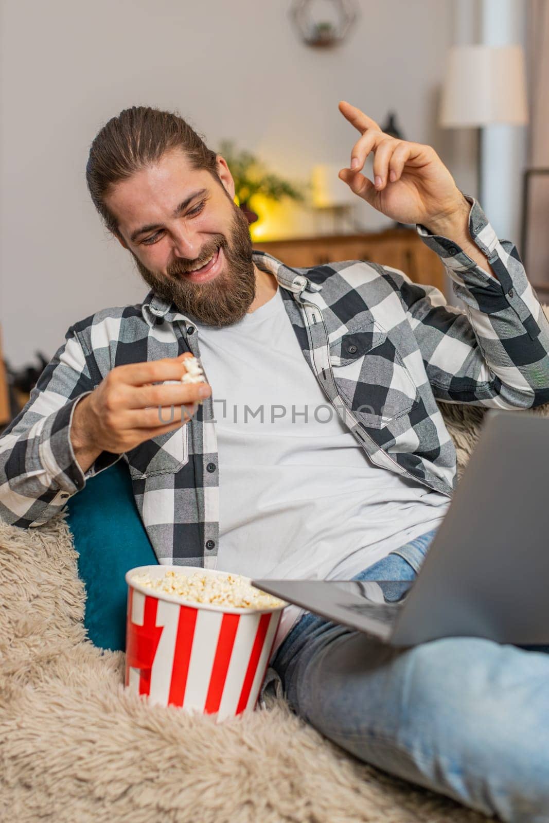 Young man eating popcorn at home, occasionally tossing kernels playfully while watching funny clips on laptop. Caucasian guy relaxes comfortably on sofa, laughing out loud at silly and amusing videos