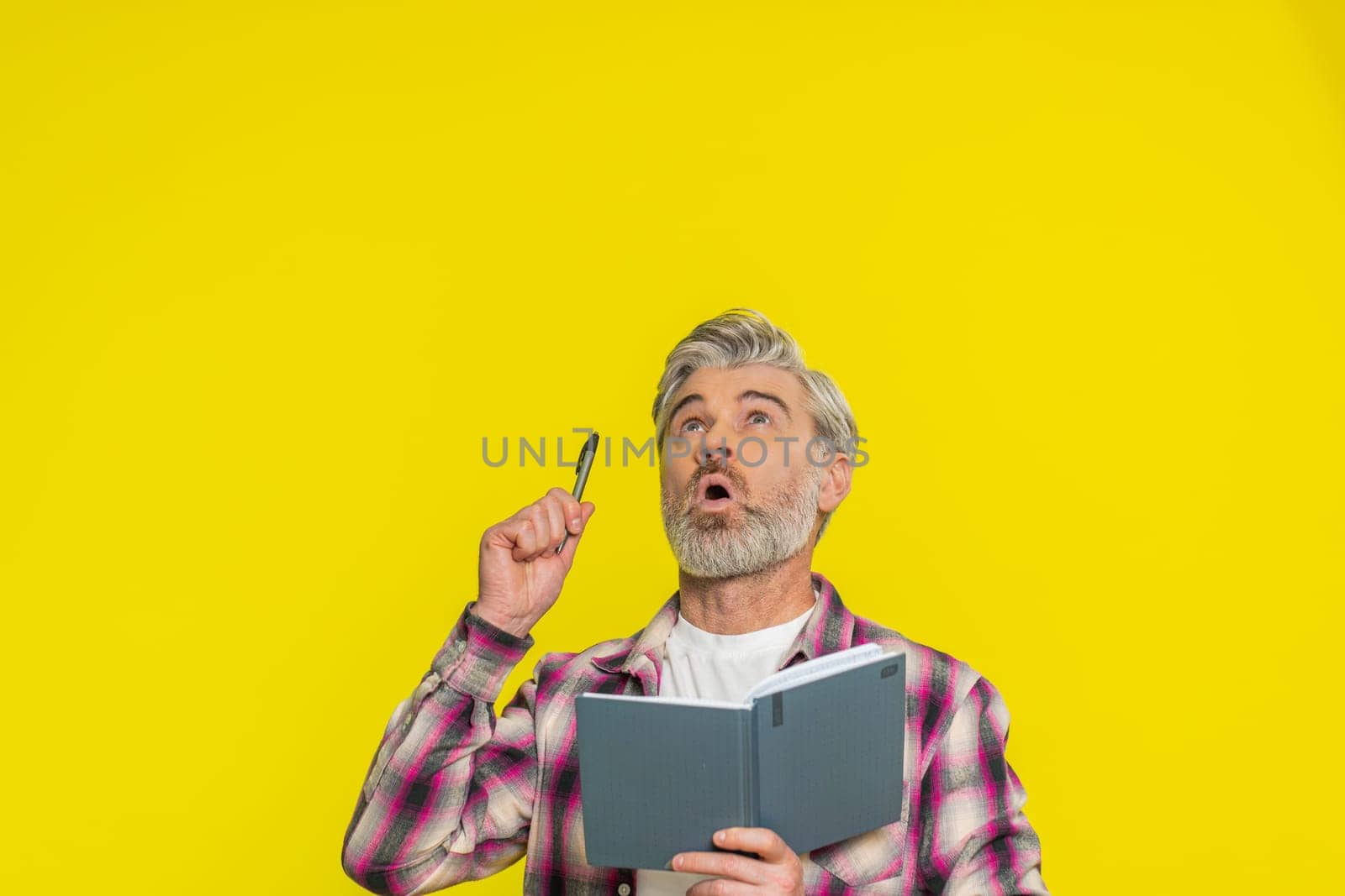 Thoughtful journalist Caucasian middle-aged man making notes, writing down thoughts with pen into notepad notebook diary, to do list, good idea. Mature old guy isolated on yellow background indoors