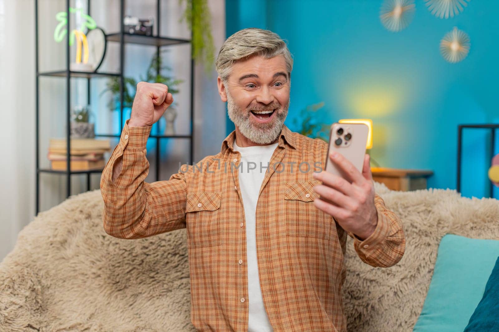 Caucasian mature man sitting on sofa at home, smiling joyfully while holding smartphone. Middle-aged guy celebrates successful project completion online game victory, expressing satisfaction happiness