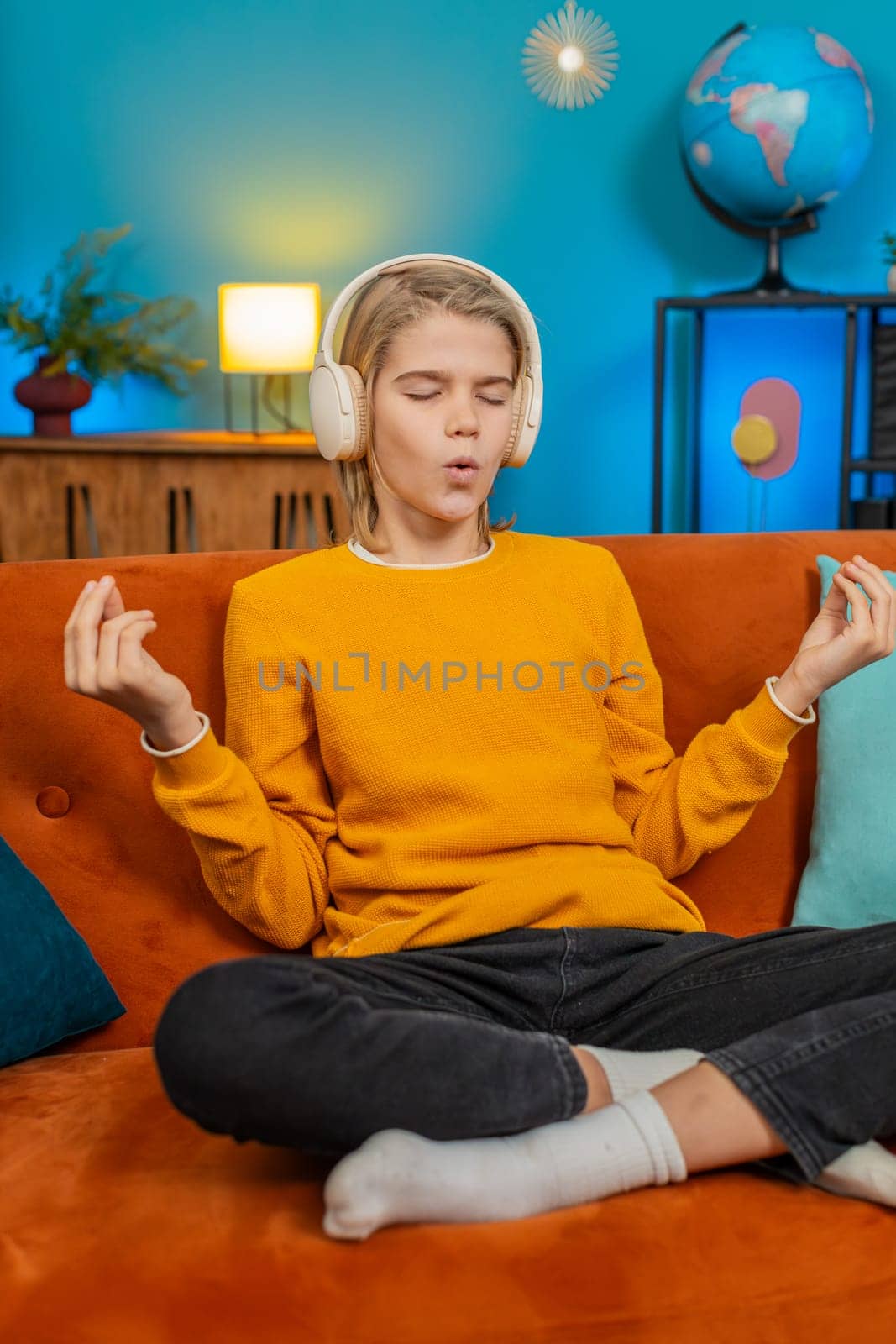 Caucasian teenager boy meditates in lotus pose on comfortable sofa at home. Wearing headphones listening music, child relaxes unwinds peacefully after long school day, finding inner peace calmness