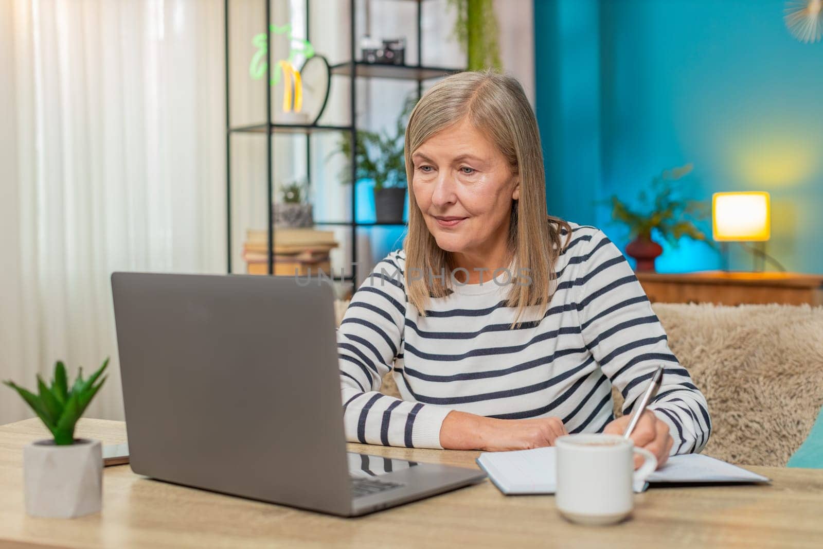 Mature woman participating in online course on laptop. Caucasian grandmother taking notes while sitting on sofa at home table, engaging in learning activities enhancing knowledge, education lessons