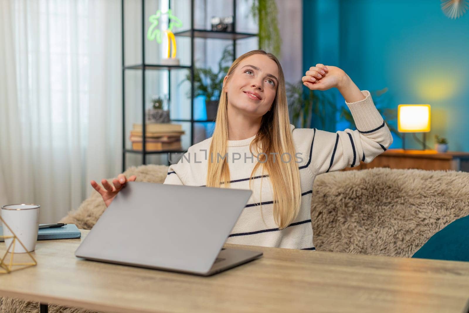 Caucasian woman sits at home on table finishing a remote workday on laptop, stretching, smiling, feeling calm and fully satisfied. Girl appears happy, relieved, enjoying calm work, continuing effort